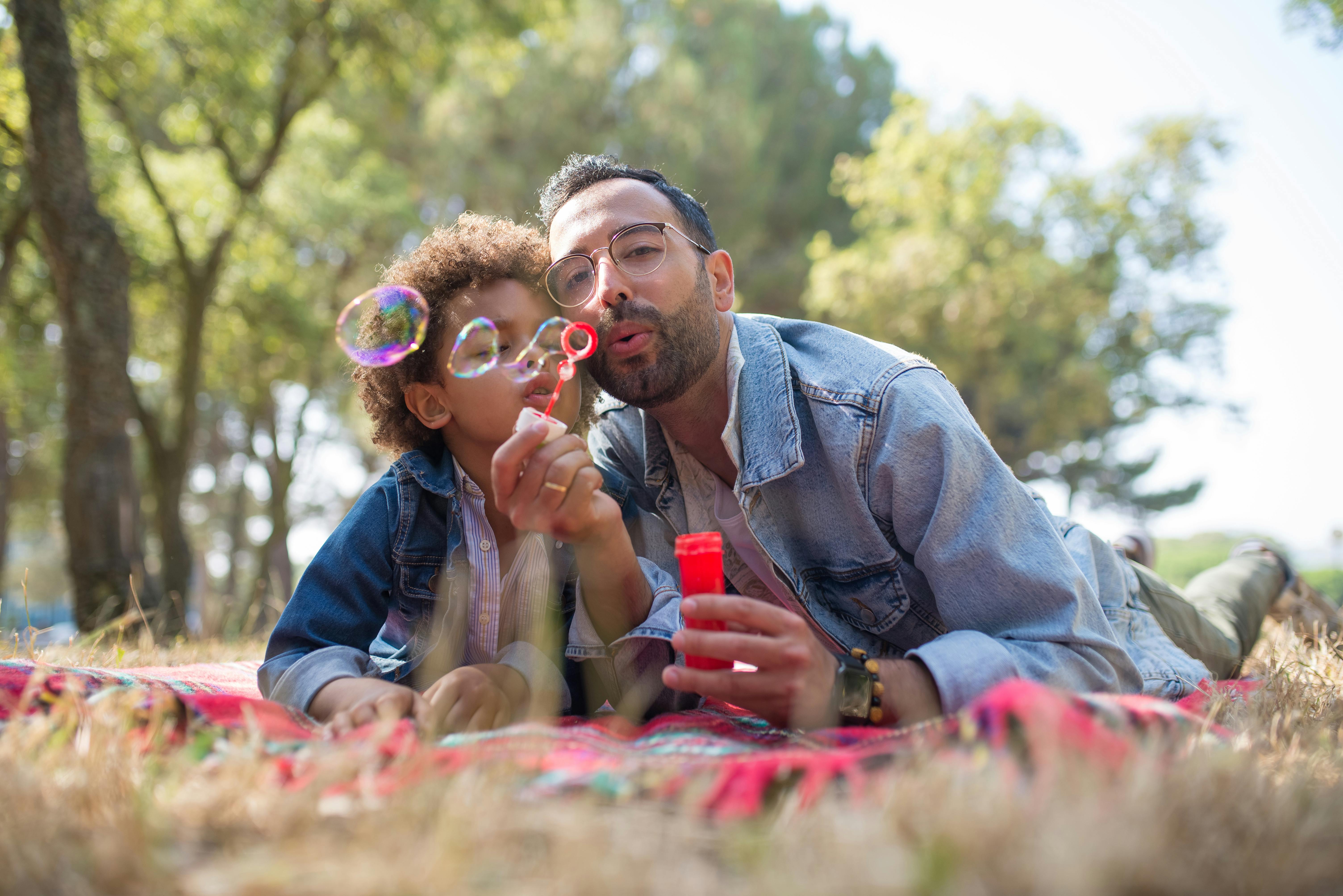 A father and sun laying down on grass and joyfully blowing bubbles together.